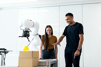 Two young engineers interacting with an AI-powered robotic arm in a modern lab, smiling as the robot picks up a yellow object near a cardboard box, showcasing the role of artificial intelligence in engineering innovation.